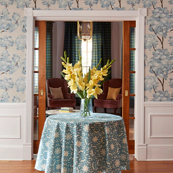 A circular table with floral table cloth and yellow flowers with in a vase. Behind is a doorway with blue floral wallpaper.
